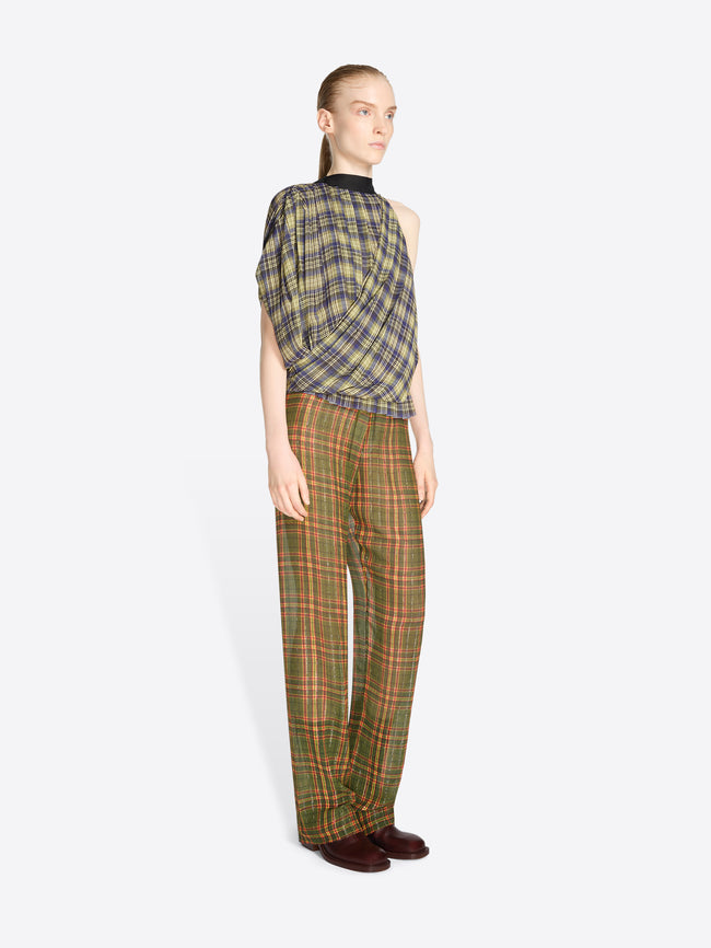 A model stands against a light gray background wearing a sleeveless asymmetrical plaid top and matching plaid trousers in olive green, brown, and beige tones, holding a patterned handbag.