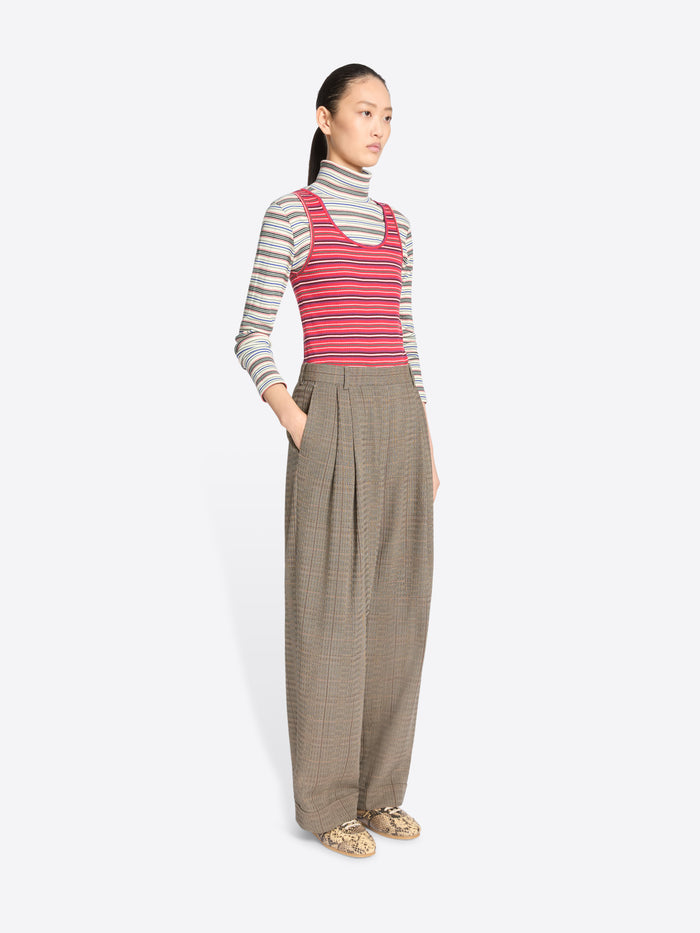 A model stands against a light gray background wearing a striped turtleneck top, a red and gray striped tank top, wide-leg pleated trousers, and snakeskin shoes.