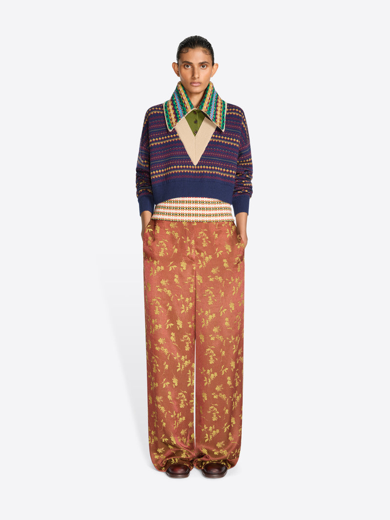 A model stands against a light gray studio background wearing a cropped knit V-neck sweater, a multicolor patterned collar and wide-leg rust-colored pants with a gold floral print.
