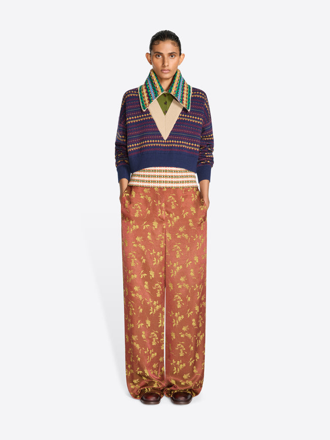A model stands against a light gray studio background wearing a cropped knit V-neck sweater, a multicolor patterned collar and wide-leg rust-colored pants with a gold floral print.