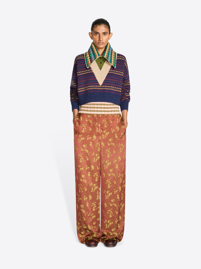 A model stands against a light gray studio background wearing a cropped knit V-neck sweater, a multicolor patterned collar and wide-leg rust-colored pants with a gold floral print.