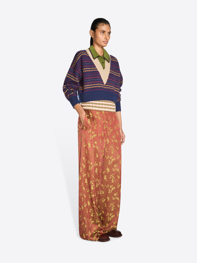 A model stands against a light gray studio background wearing a cropped knit V-neck sweater, a multicolor patterned collar and wide-leg rust-colored pants with a gold floral print.