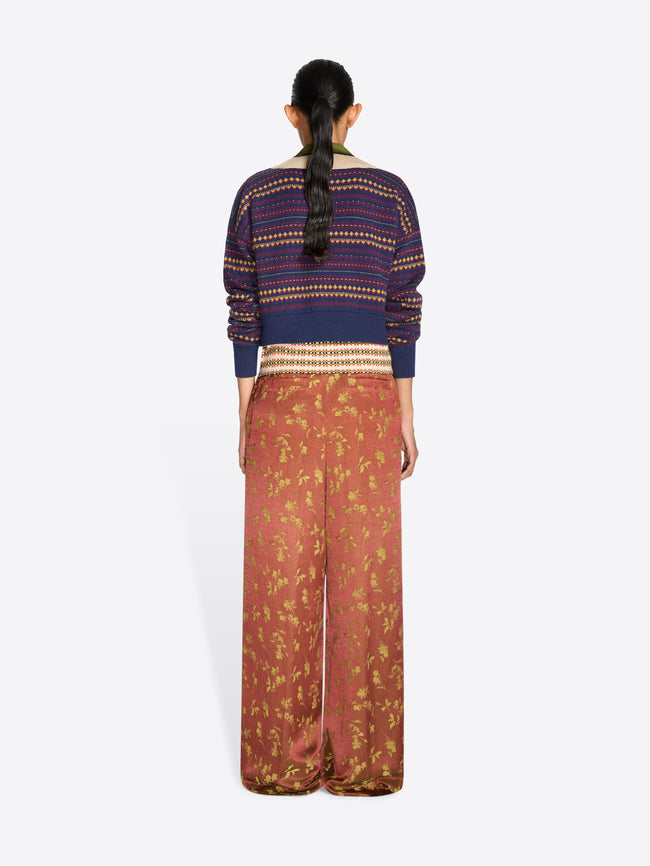 A model stands against a light gray studio background wearing a cropped knit V-neck sweater, a multicolor patterned collar and wide-leg rust-colored pants with a gold floral print.