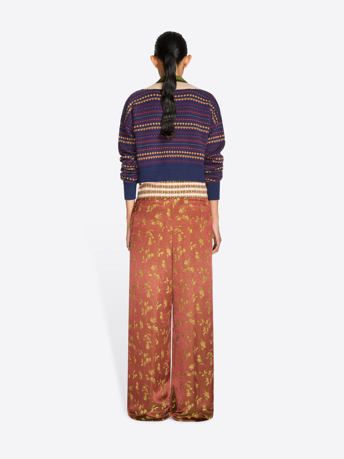 A model stands against a light gray studio background wearing a cropped knit V-neck sweater, a multicolor patterned collar and wide-leg rust-colored pants with a gold floral print.