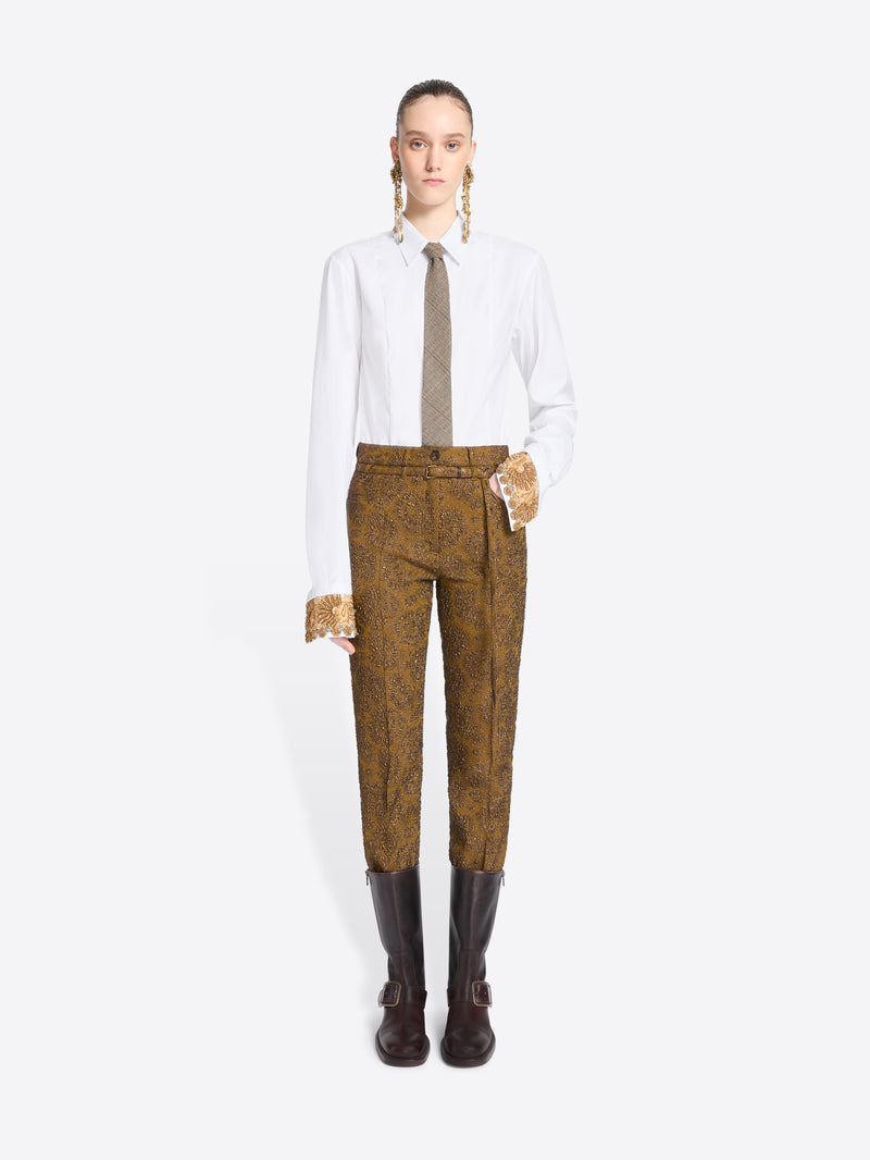 A model stands against a light gray background wearing a white long-sleeve shirt, a taupe tie, mustard-brown patterned pants, and brown boots.