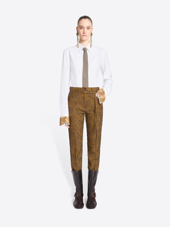 A model stands against a light gray background wearing a white long-sleeve shirt, a taupe tie, mustard-brown patterned pants, and brown boots.