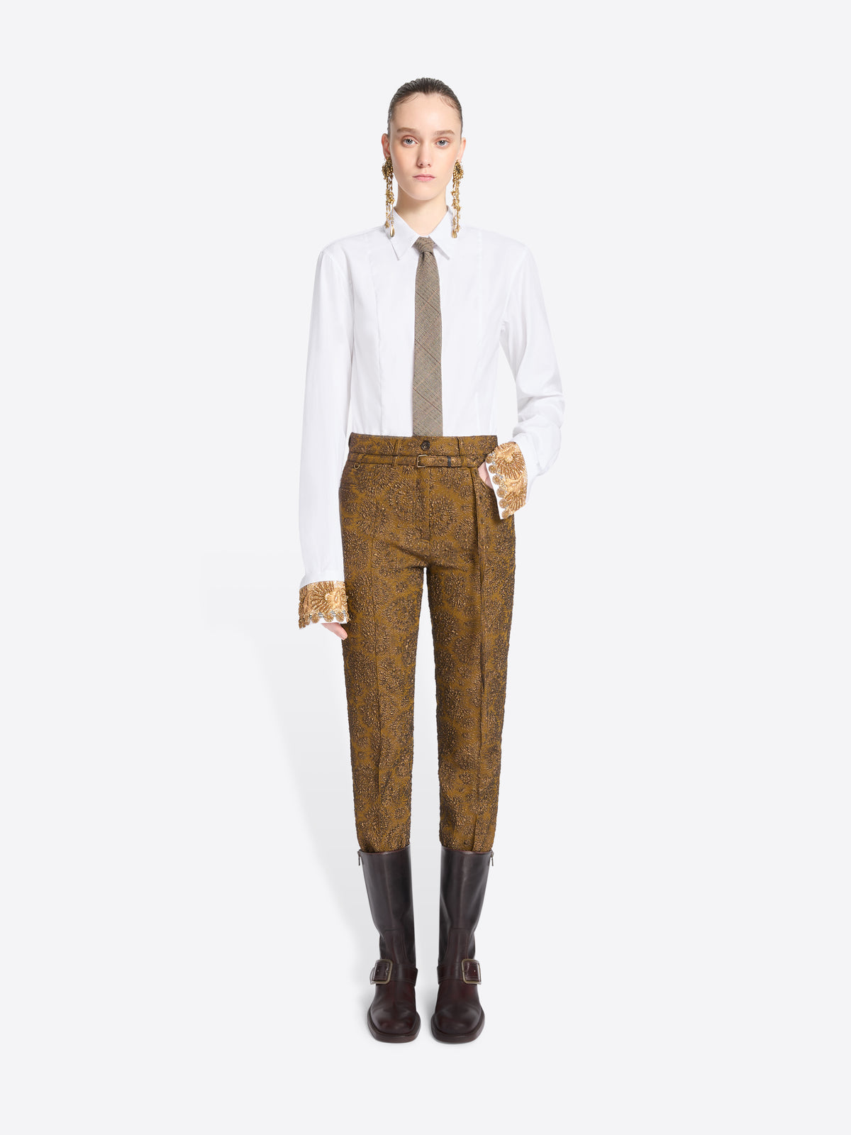 A model stands against a light gray background wearing a white long-sleeve shirt, a taupe tie, mustard-brown patterned pants, and brown boots.