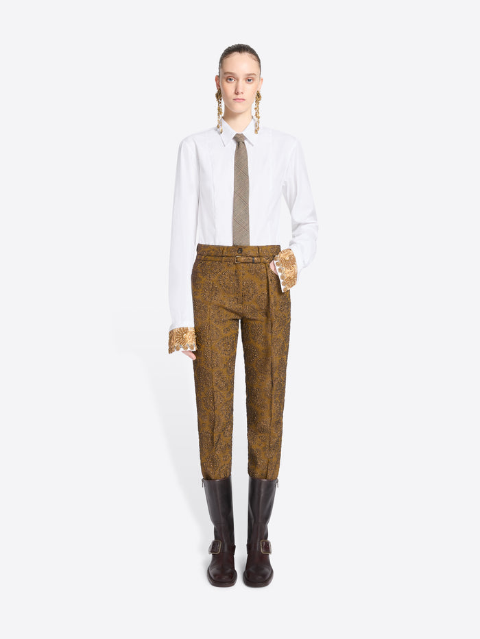 A model stands against a light gray background wearing a white long-sleeve shirt, a taupe tie, mustard-brown patterned pants, and brown boots.