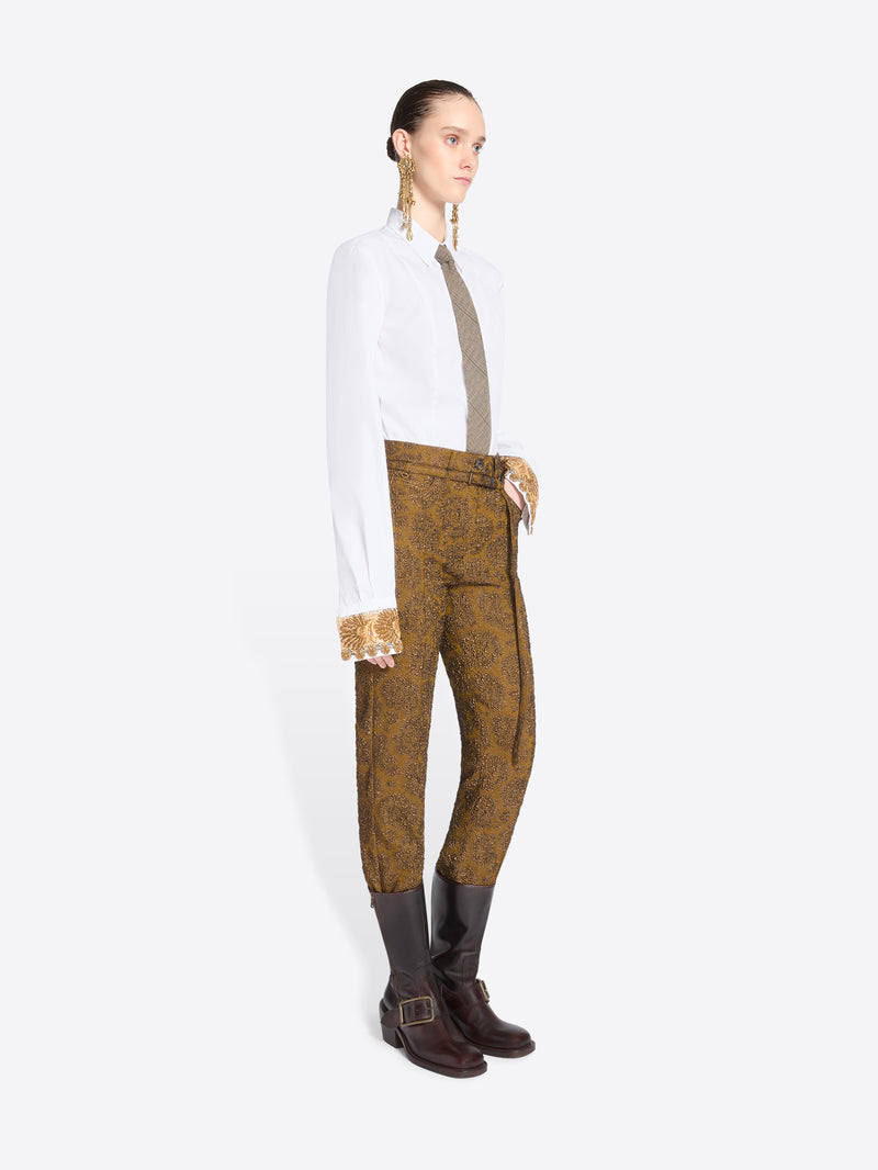 A model stands against a light gray background wearing a white long-sleeve shirt, a taupe tie, mustard-brown patterned pants, and brown boots.