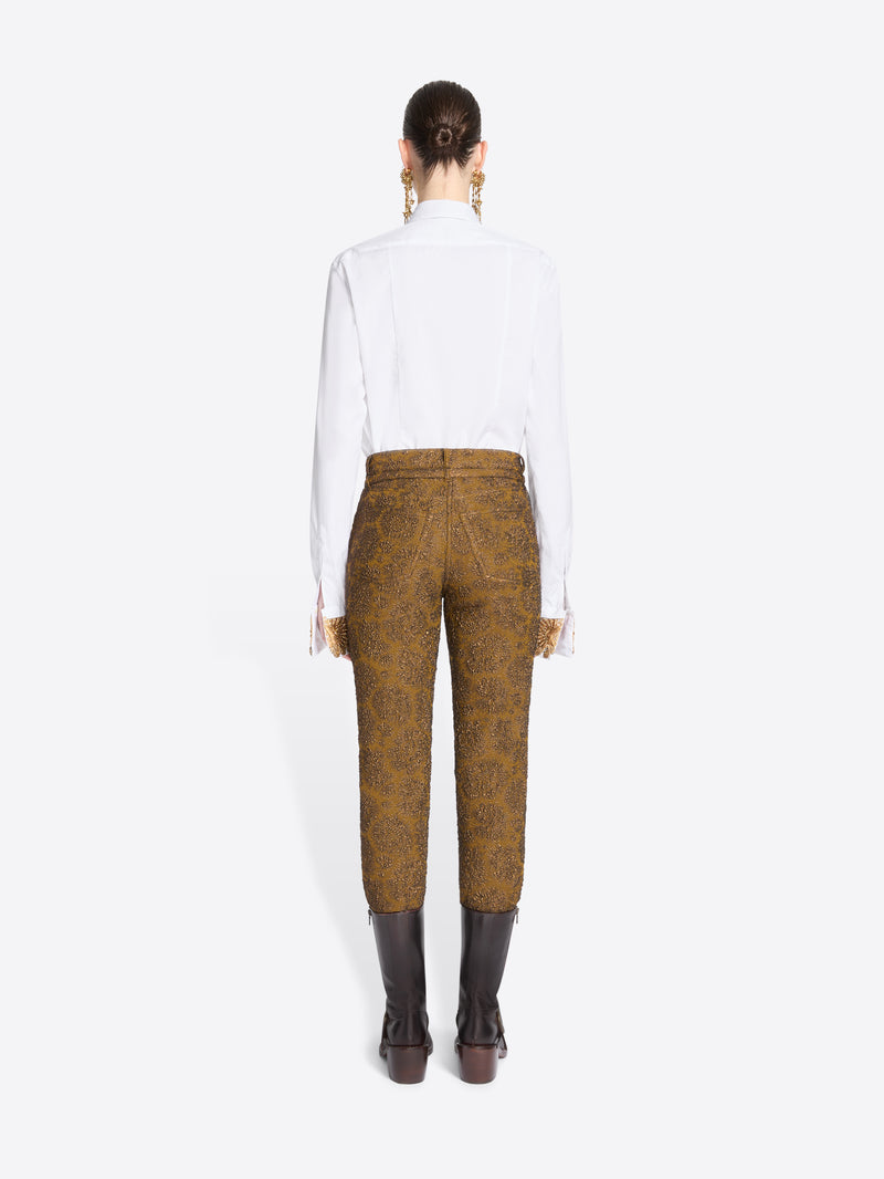 Back view of a model standing against a light gray background wearing a white long-sleeve shirt, a taupe tie, mustard-brown patterned pants, and brown boots.