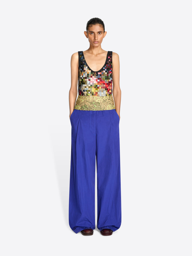 A model stands against a light gray background wearing a sleeveless top with a multicolor pixelated pattern and a gold textured waistband, paired with wide-leg royal blue pants.