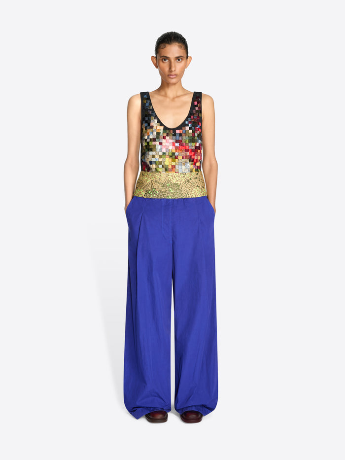 A model stands against a light gray background wearing a sleeveless top with a multicolor pixelated pattern and a gold textured waistband, paired with wide-leg royal blue pants.