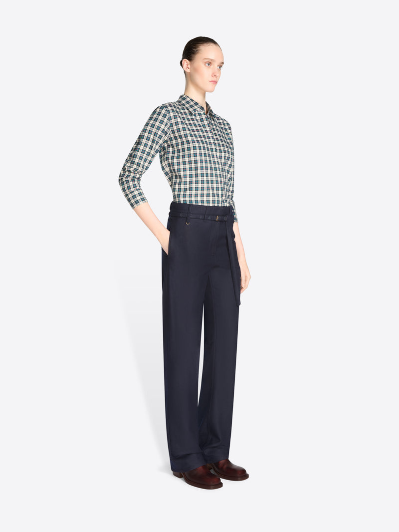 A model stands against a light gray background wearing a green and white checkered button-down shirt, dark navy pants, and brown shoes, while holding a large orange and multicolor patterned tote bag.