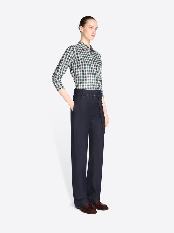 A model stands against a light gray background wearing a green and white checkered button-down shirt, dark navy pants, and brown shoes, while holding a large orange and multicolor patterned tote bag.