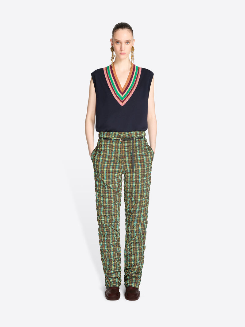 A model stands against a light gray background wearing a sleeveless navy sweater vest with V-neck stripe trim, paired with green and black plaid pants.