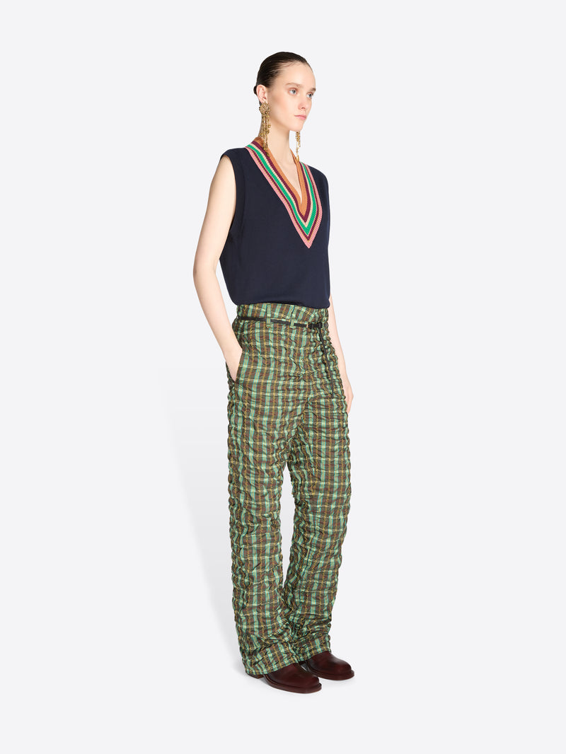 A model stands against a light gray background wearing a sleeveless navy sweater vest with V-neck stripe trim, paired with green and black plaid pants.