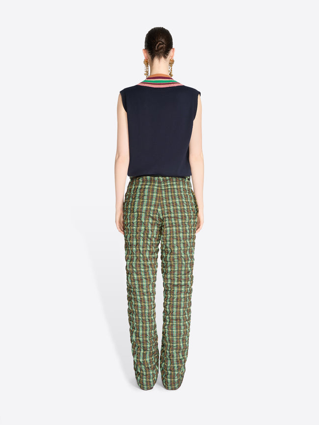 Back view of a model standing against a light gray background wearing a sleeveless navy sweater vest with V-neck stripe trim, paired with green and black plaid pants.