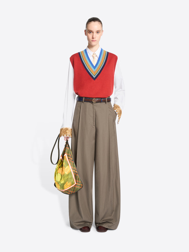 A model stands against a light gray background wearing a red knit vest with blue, yellow, and green striped trim over a white collared shirt, paired with wide-leg taupe trousers and holding a patterned handbag.