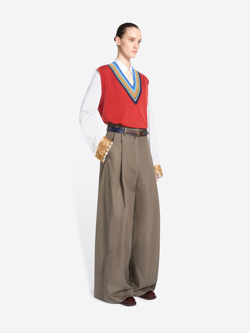 A model stands against a light gray background wearing a red knit vest with blue, yellow, and green striped trim over a white collared shirt, paired with wide-leg taupe trousers. 