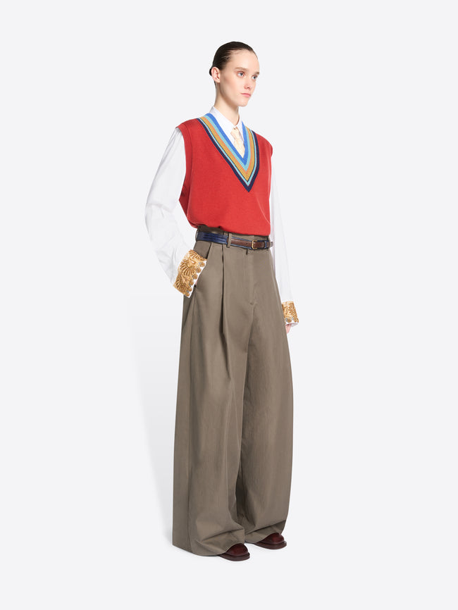 A model stands against a light gray background wearing a red knit vest with blue, yellow, and green striped trim over a white collared shirt, paired with wide-leg taupe trousers. 