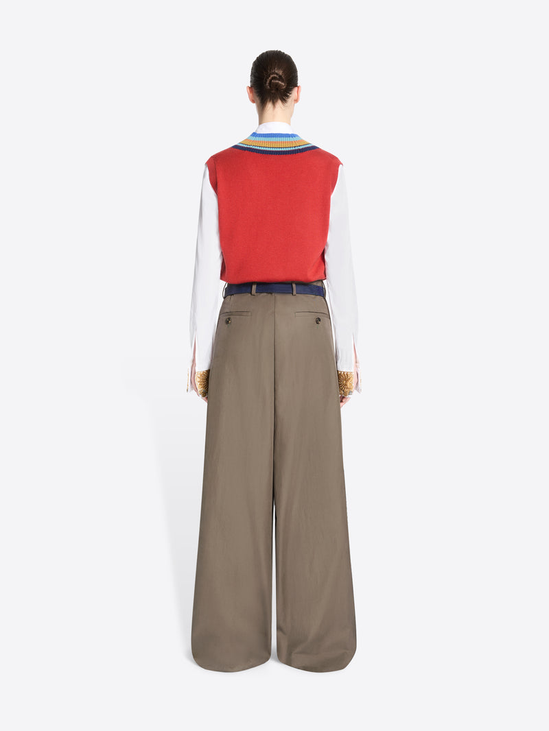 Back view of a model standing against a light gray background wearing a red knit vest with blue, yellow, and green striped trim over a white collared shirt, paired with wide-leg taupe trousers and holding a patterned handbag.