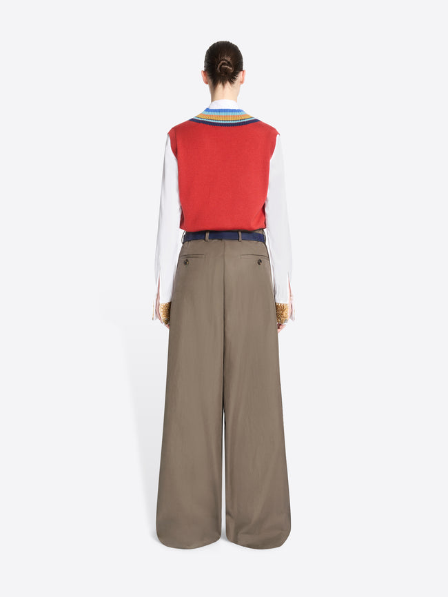 Back view of a model standing against a light gray background wearing a red knit vest with blue, yellow, and green striped trim over a white collared shirt, paired with wide-leg taupe trousers and holding a patterned handbag.