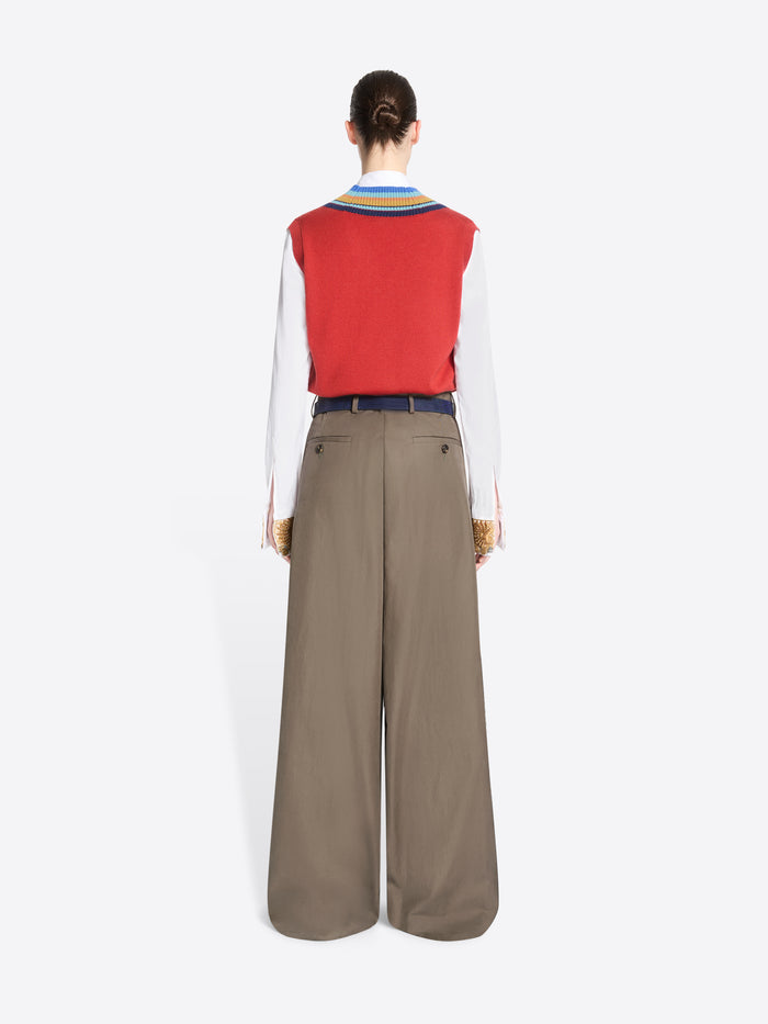 Back view of a model standing against a light gray background wearing a red knit vest with blue, yellow, and green striped trim over a white collared shirt, paired with wide-leg taupe trousers and holding a patterned handbag.