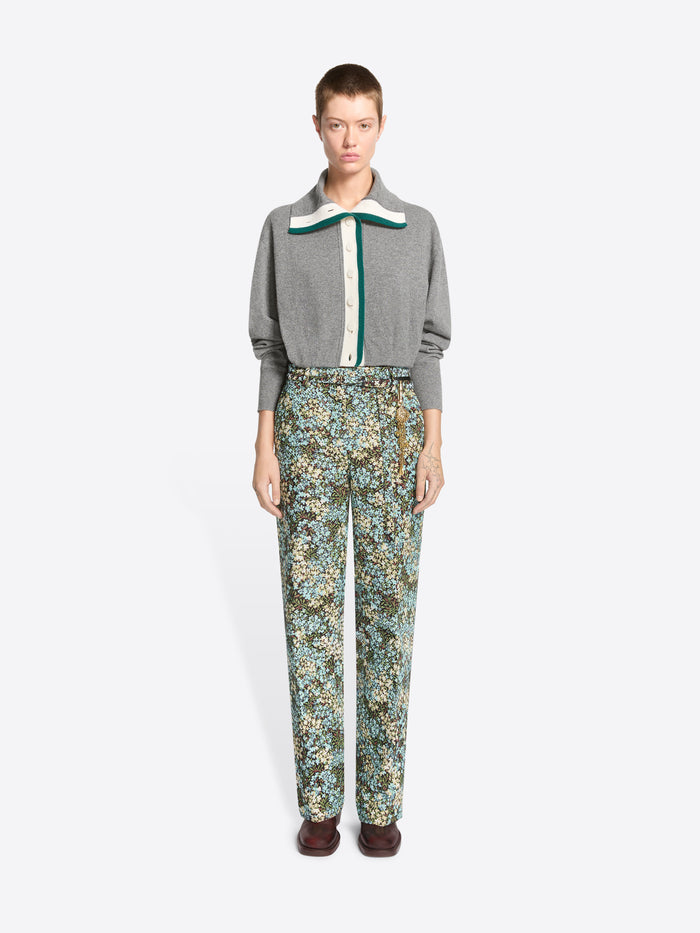 A model stands against a light gray background wearing a gray knit cardigan with a green and white collar detail, paired with high-waisted floral patterned trousers and dark brown shoes.
