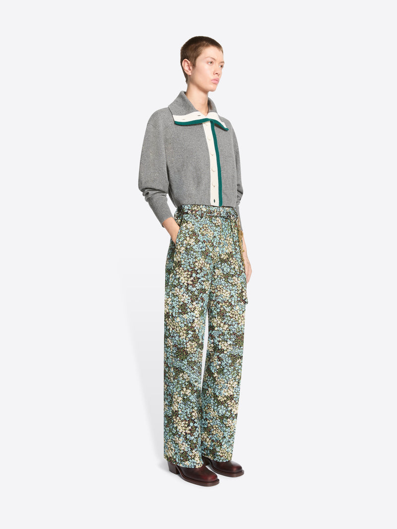 A model stands against a light gray background wearing a gray knit cardigan with a green and white collar detail, paired with high-waisted floral patterned trousers and dark brown shoes.