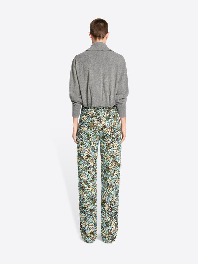 Back view of a model standing against a light gray background wearing a gray knit cardigan paired with high-waisted floral patterned trousers.