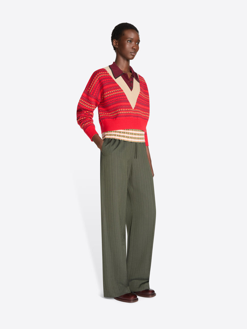 A model stands against a light gray background wearing a red and beige patterned knit sweater over a burgundy collared shirt, paired with olive green wide-leg trousers.