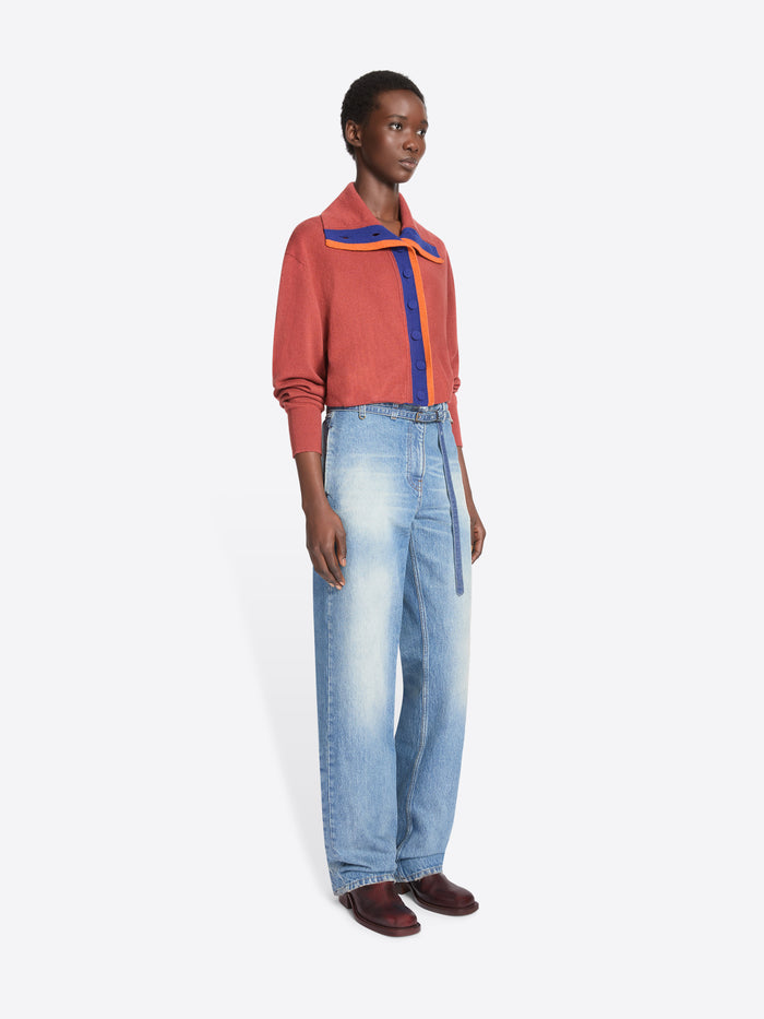 A model stands against a light gray background wearing a long-sleeve coral cardigan with a purple center placket, and a wide collar with purple and orange accents, paired with light-wash wide-leg jeans and dark brown shoes.
