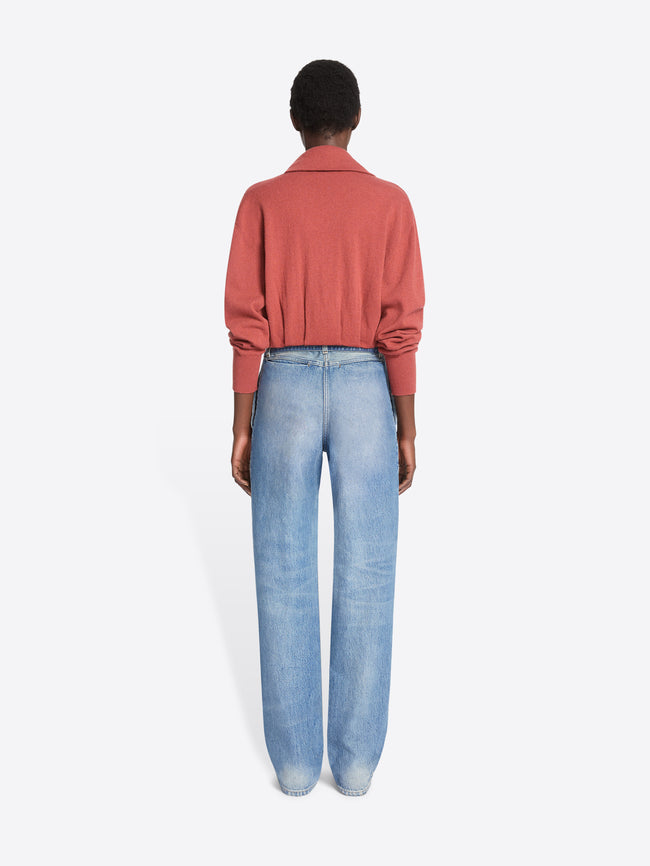 Back view of a model stands against a light gray background wearing a long-sleeve coral cardigan, paired with light-wash wide-leg jeans.