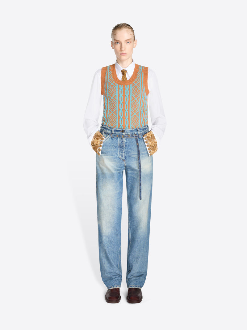 A model stands against a light gray background wearing a white long-sleeve shirt, a patterned orange and blue knit vest, light blue jeans with belt at the waist and dark brown shoes.