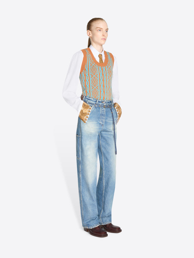 A model stands against a light gray background wearing a white long-sleeve shirt, a patterned orange and blue knit vest, light blue jeans with belt at the waist and dark brown shoes.