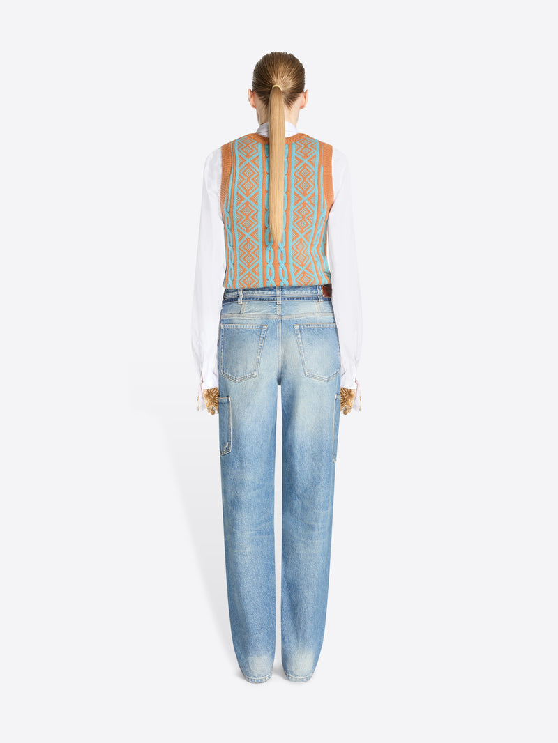 A back view of model standing against a light gray background wearing a white long-sleeve shirt, a patterned knit vest and light blue jeans with belt. 