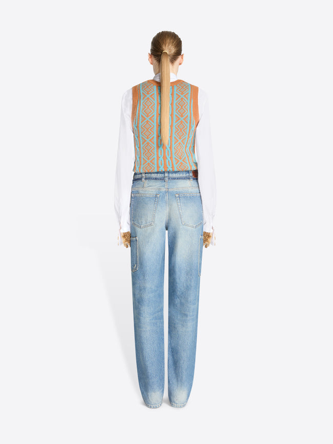 A back view of model standing against a light gray background wearing a white long-sleeve shirt, a patterned knit vest and light blue jeans with belt. 