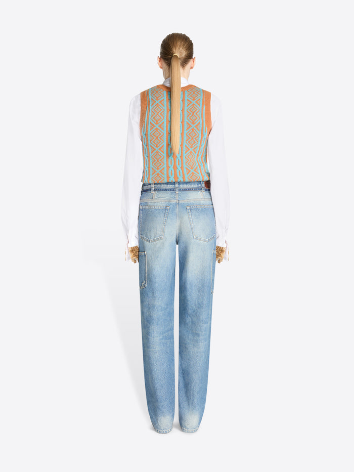 A back view of model standing against a light gray background wearing a white long-sleeve shirt, a patterned knit vest and light blue jeans with belt. 