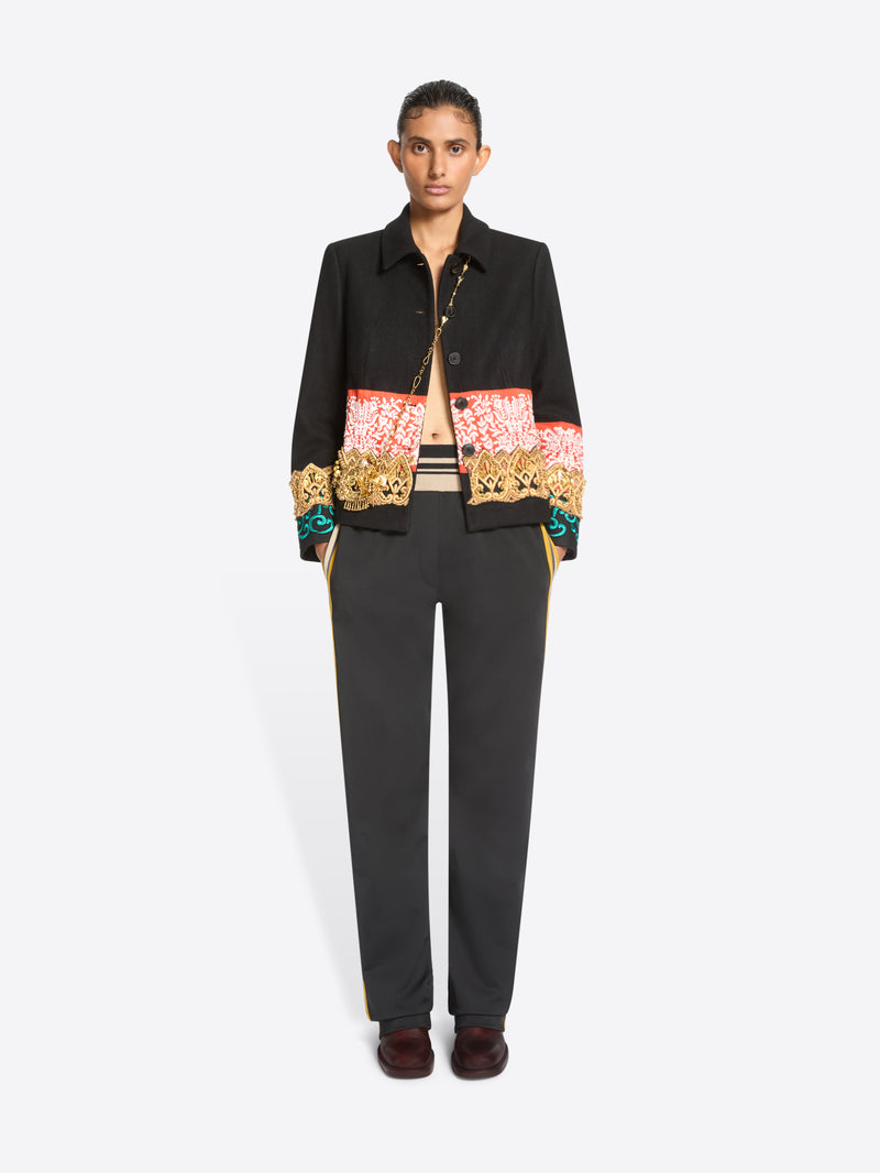 A model stands against a light gray background wearing a black cropped jacket with ornate multicolor embroidered detailing across the chest and cuffs, paired with dark charcoal pants with yellow side stripes and brown shoes.