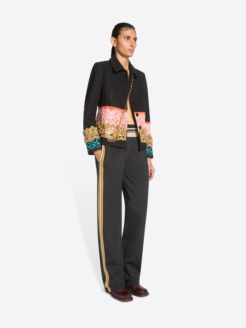 A model stands against a light gray background wearing a black cropped jacket with ornate multicolor embroidered detailing across the chest and cuffs, paired with dark charcoal pants with yellow side stripes and brown shoes.