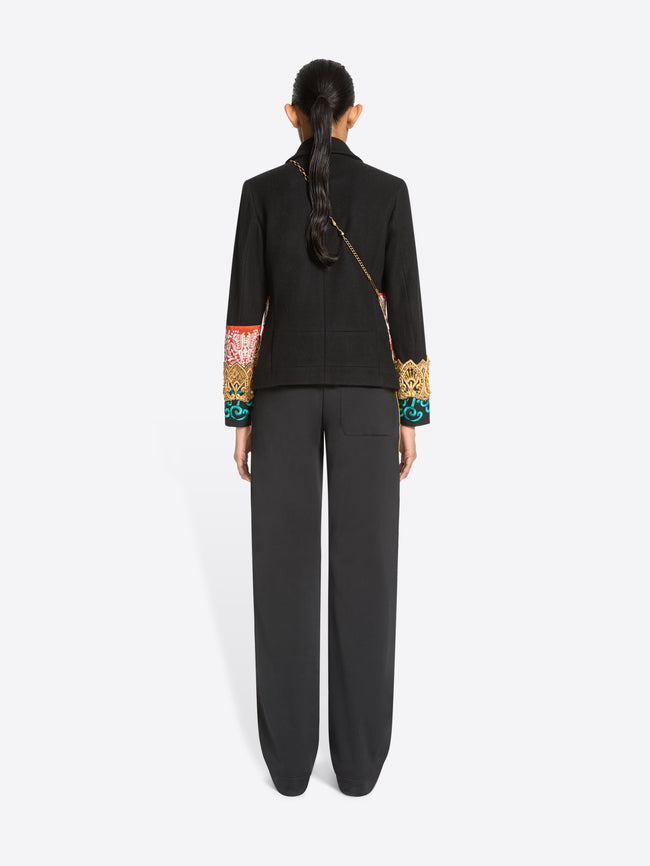 Back view of a model standing against a light gray background wearing a black cropped jacket with ornate multicolor embroidered detailing across the chest and cuffs, paired with dark charcoal pants.