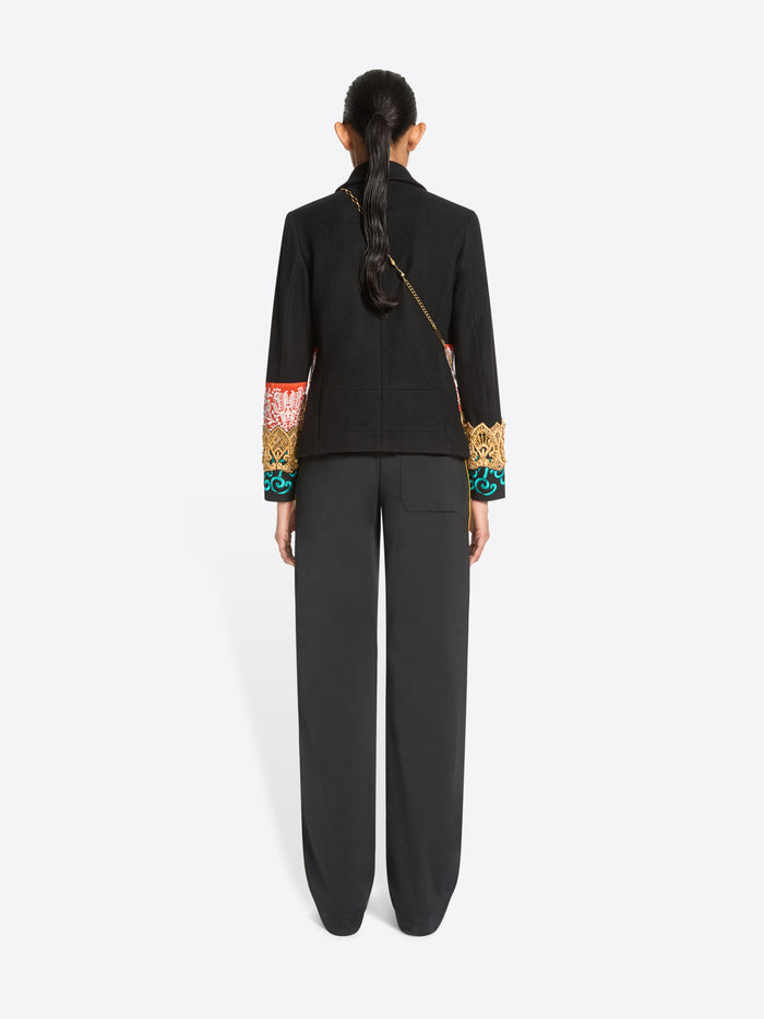 Back view of a model standing against a light gray background wearing a black cropped jacket with ornate multicolor embroidered detailing across the chest and cuffs, paired with dark charcoal pants.
