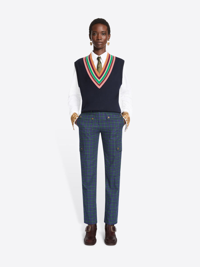 A model stands against a light gray background wearing a navy sweater vest with a colorful striped V-neck trim over a white collared shirt and tie, paired with navy plaid trousers and dark brown shoes.