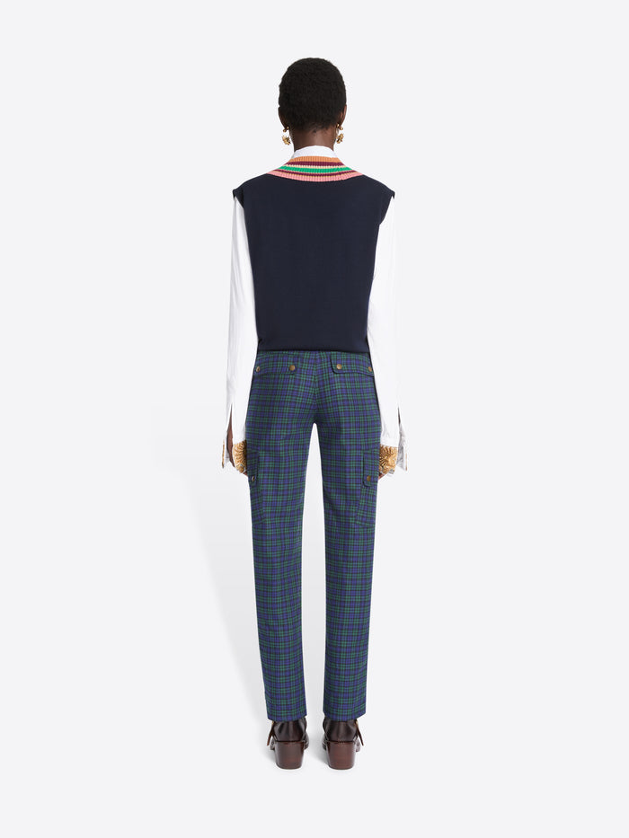 Back view of a model standing against a light gray background wearing a navy sweater vest with a colorful striped V-neck trim over a white collared shirt, paired with navy plaid trousers and dark brown shoes.