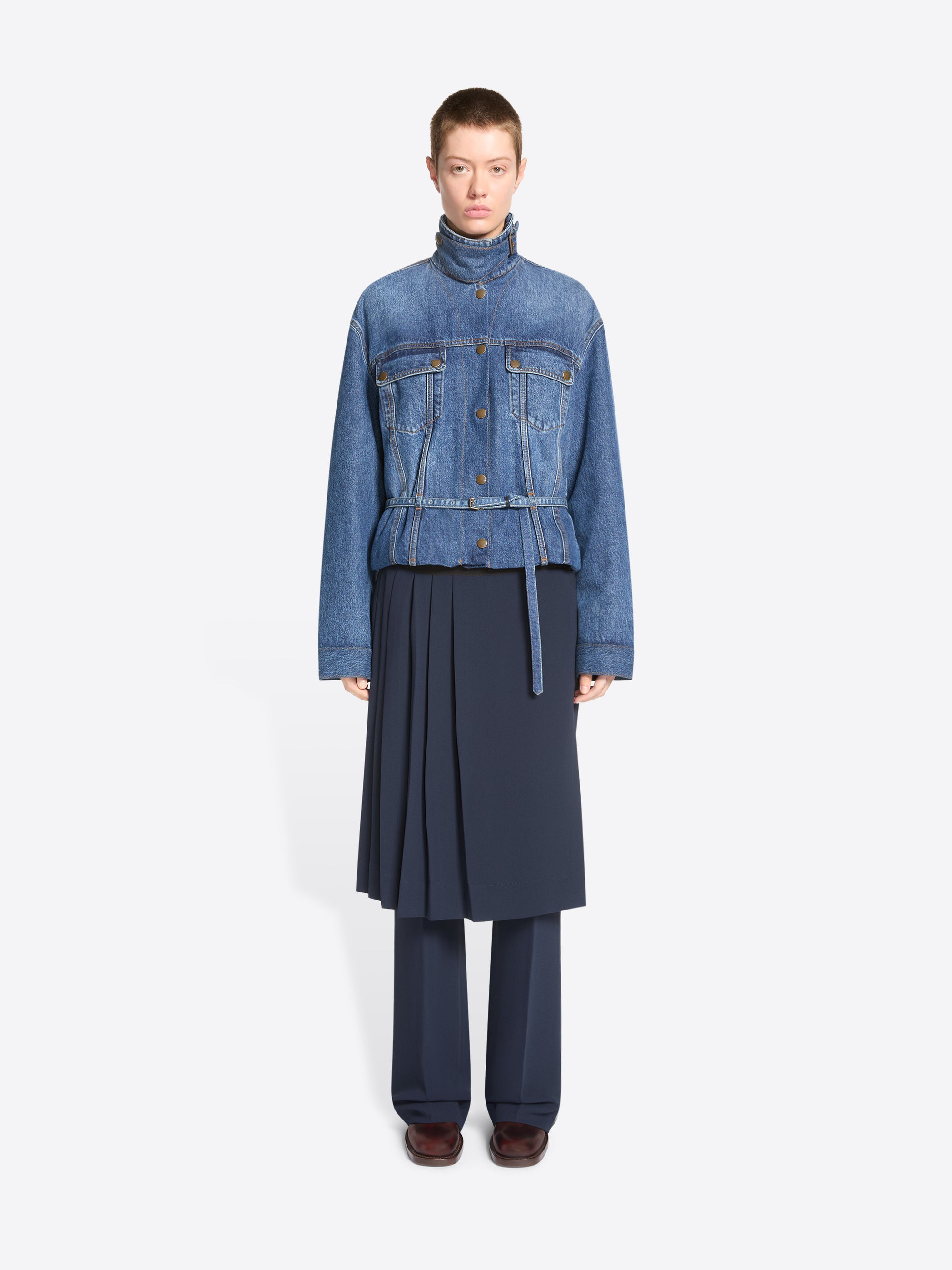 A model stands against a light gray background wearing a cropped blue denim jacket with a high mock neck, button front, and flap chest pockets, paired with a dark navy pleated skirt and matching navy trousers.