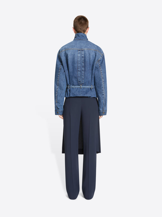 Back view of a model standing against a light gray background wearing a cropped blue denim jacket with a high mock neck, paired with a dark navy pleated skirt and matching navy trousers.