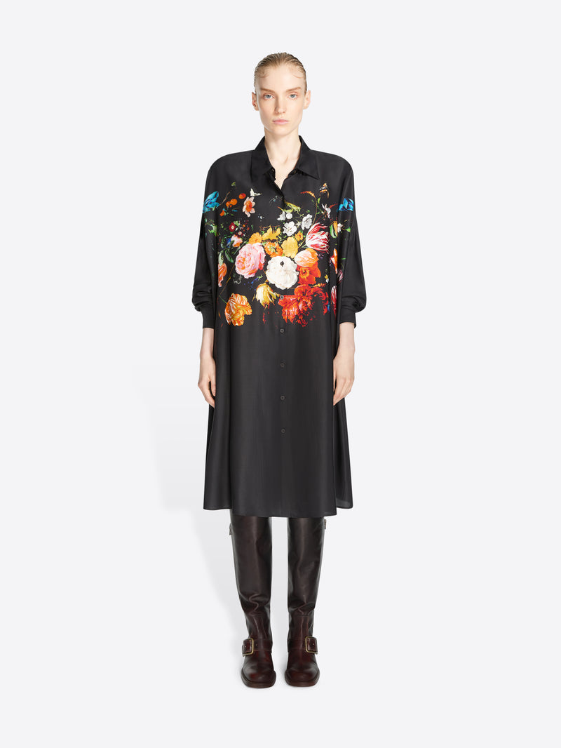A model stands against a light gray background wearing a black long-sleeve shirt dress with a large multicolor floral print across the chest and shoulders, paired with dark brown knee-high boots.