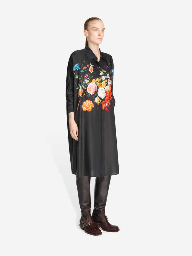 A model stands against a light gray background wearing a black long-sleeve shirt dress with a large multicolor floral print across the chest and shoulders, paired with dark brown knee-high boots.