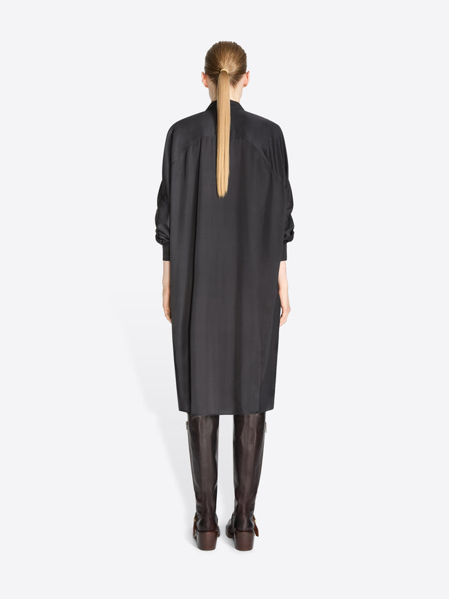 A back view of a model standing against a light gray background wearing a black long-sleeve shirt dress paired with dark brown knee-high boots.