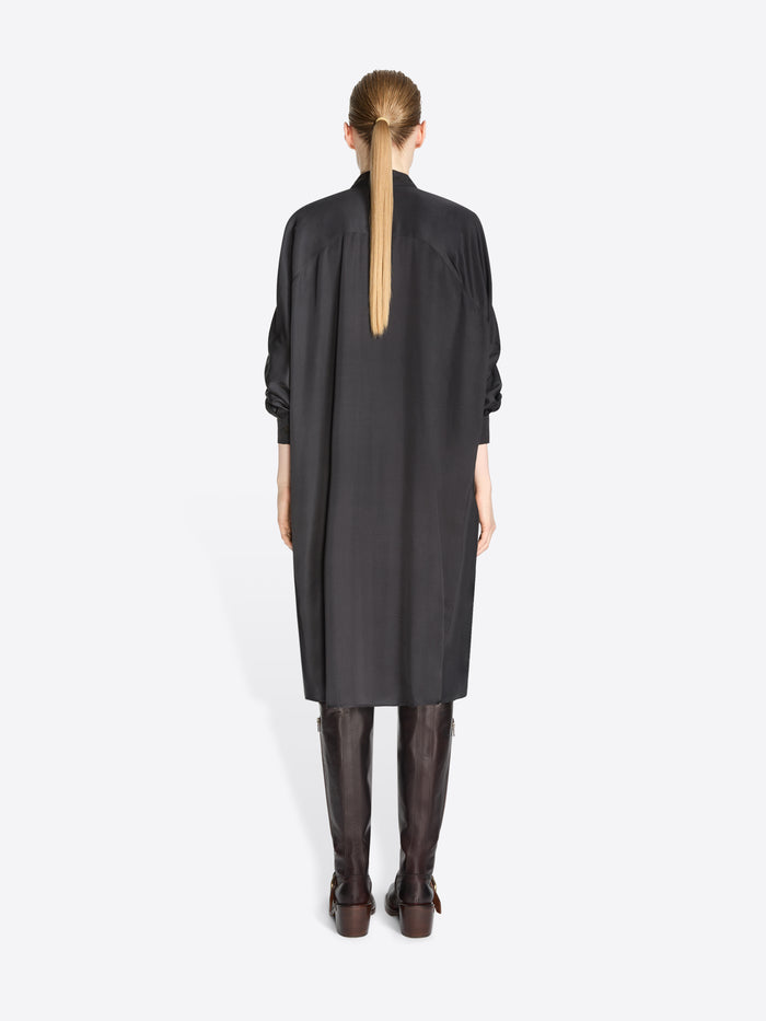 A back view of a model standing against a light gray background wearing a black long-sleeve shirt dress paired with dark brown knee-high boots.